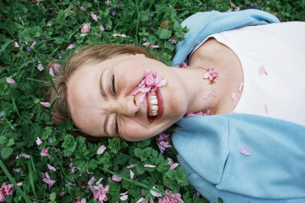A happy woman lies on grass, smiling with pink flower petals around her face.