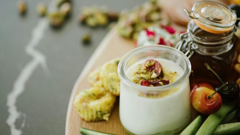 High angle of delicious yogurt with decorative ingredients in glass near cucumber slices and cherries near jar with honey on table in light room