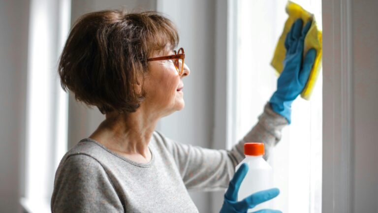 Elderly woman cleansing window indoors using gloves and cleaner.