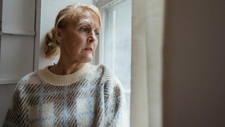 A senior woman in deep thought gazing out a window indoors.