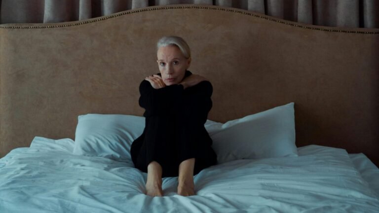 A senior woman sits alone on a bed, expressing introspection and melancholy.